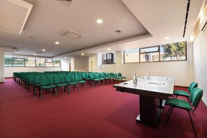 Un salón de conferencias con filas de sillas verdes, una mesa grande y ventanas amplias.