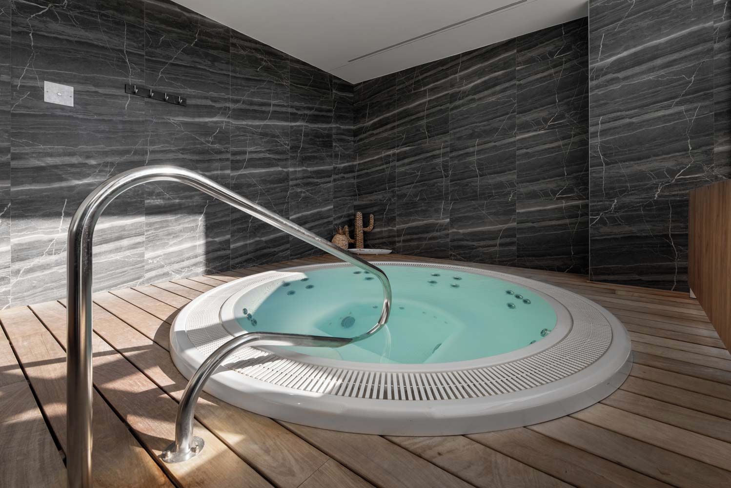 Banheira de hidromassagem moderna em ambiente interno, com deck de madeira, em frente a paredes de mármore escuro.