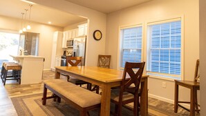 A modern kitchen with white cabinets, a wooden dining table, and a bench. There are two windows with blinds, a clock on the wall, and a kitchen island with stools. - Harbor Pointe - Modern, Close to Lake Michigan! (South Haven)