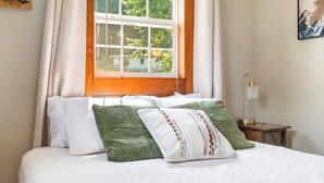 A neatly made bed with white bedding and green and white pillows, a window with curtains, and a bedside table with a lamp. - Fishing · Kayaks · Firepit · Paddleboat (Pierceton)