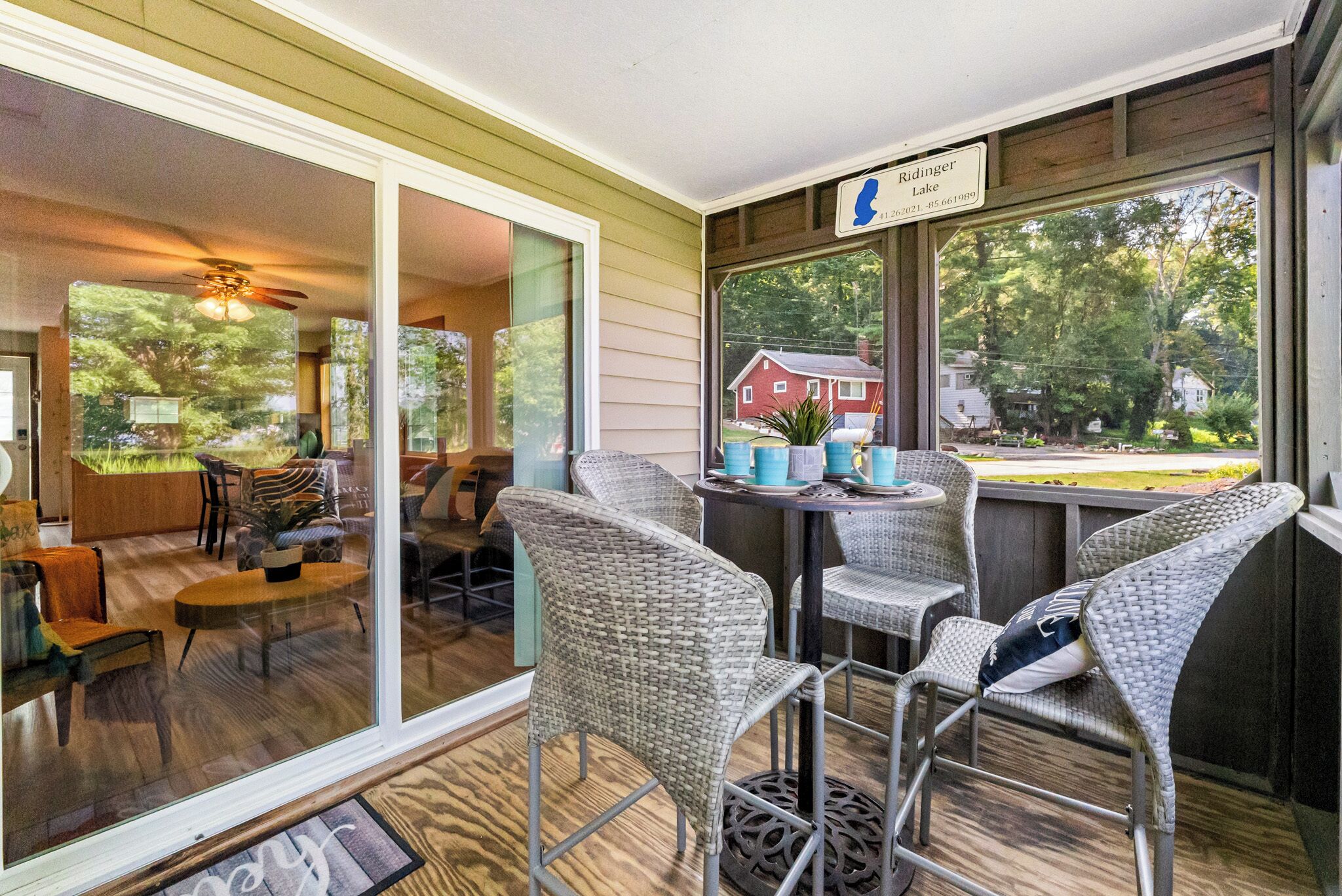 A covered outdoor seating area with wicker chairs and a table, overlooking a residential street and a lake.