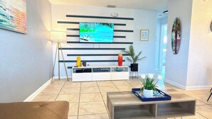 A modern living room with a flat-screen TV mounted on a wall, a wooden coffee table, a sofa, and a potted plant. - Family and pet friendly ~ Pool, gym, games! (Indian Harbour Beach)