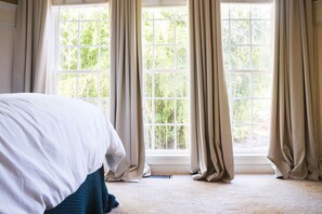 A bedroom with a large bed, beige curtains, and a view of greenery through the window. - Classic City Retreat in Five Points, Athens GA | Theater | Hot Tub | Christmas (Athens)