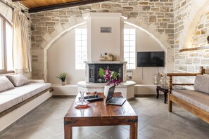 Un salon chaleureux avec une cheminée en pierre, un canapé, une table basse en bois sur laquelle se trouve un ordinateur portable, et un téléviseur fixé au mur.