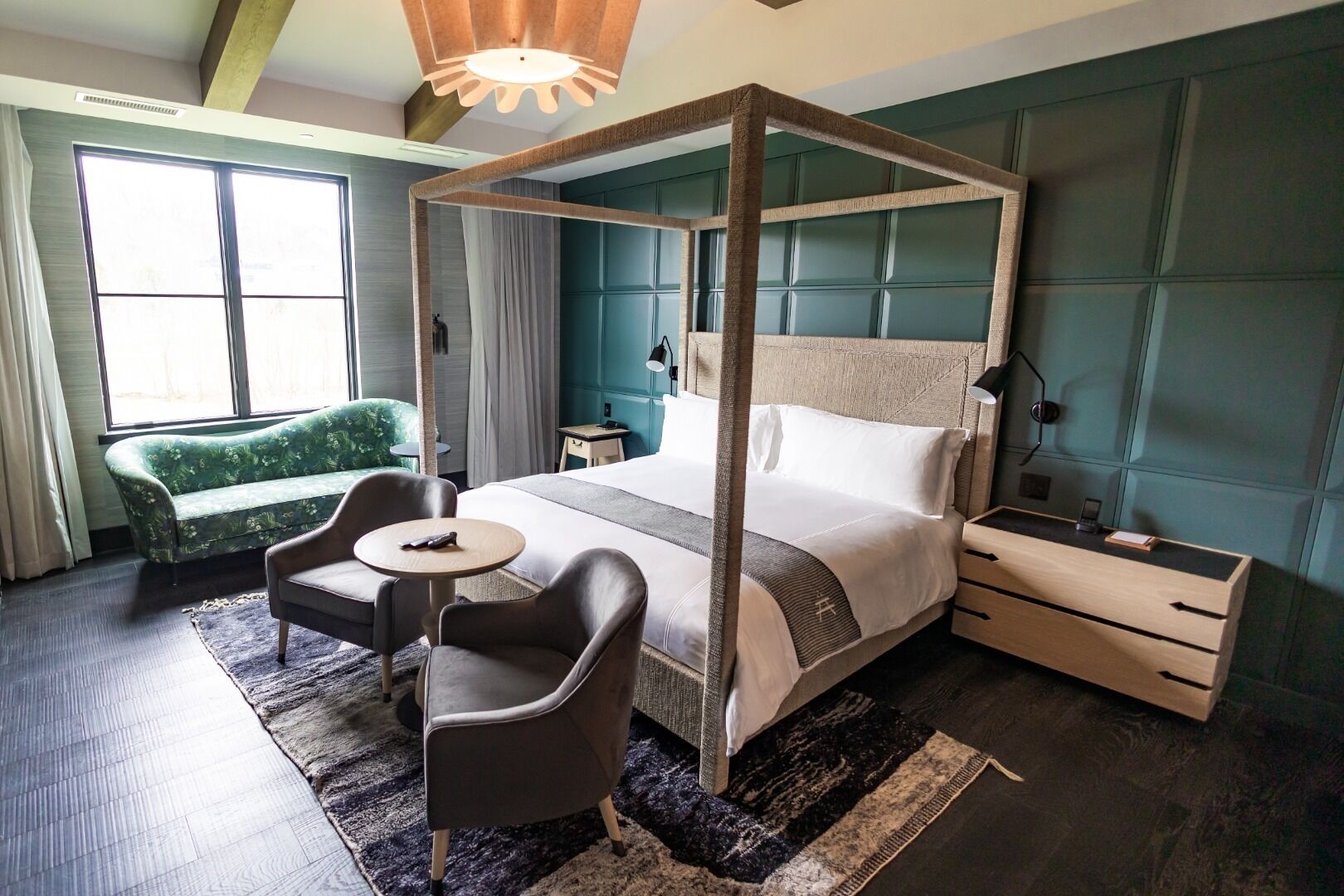 A four-poster bed with a wooden frame, a green upholstered sofa, a small round table, and a bedside table with a lamp.