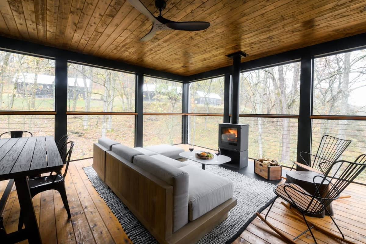 A cozy room with a wooden ceiling, large windows, a sofa, a table, and a wood-burning stove.