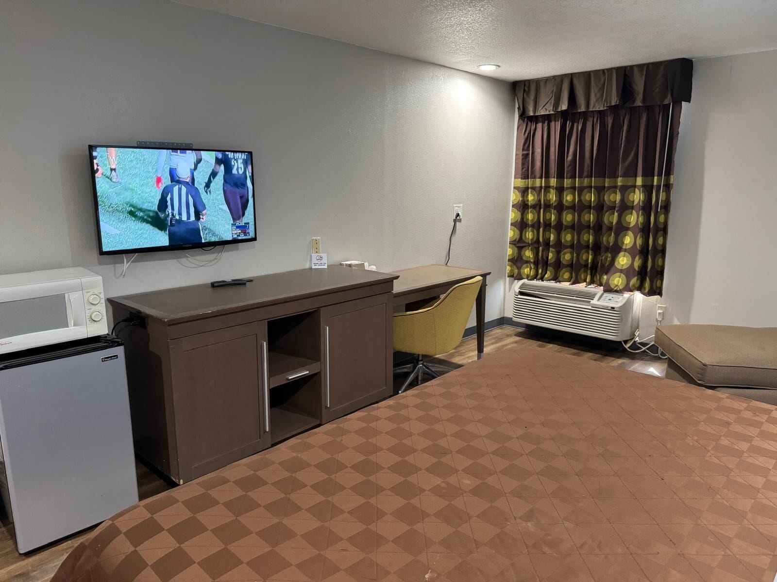 A hotel room with a bed, a desk, a television, a microwave, and an air conditioning unit.