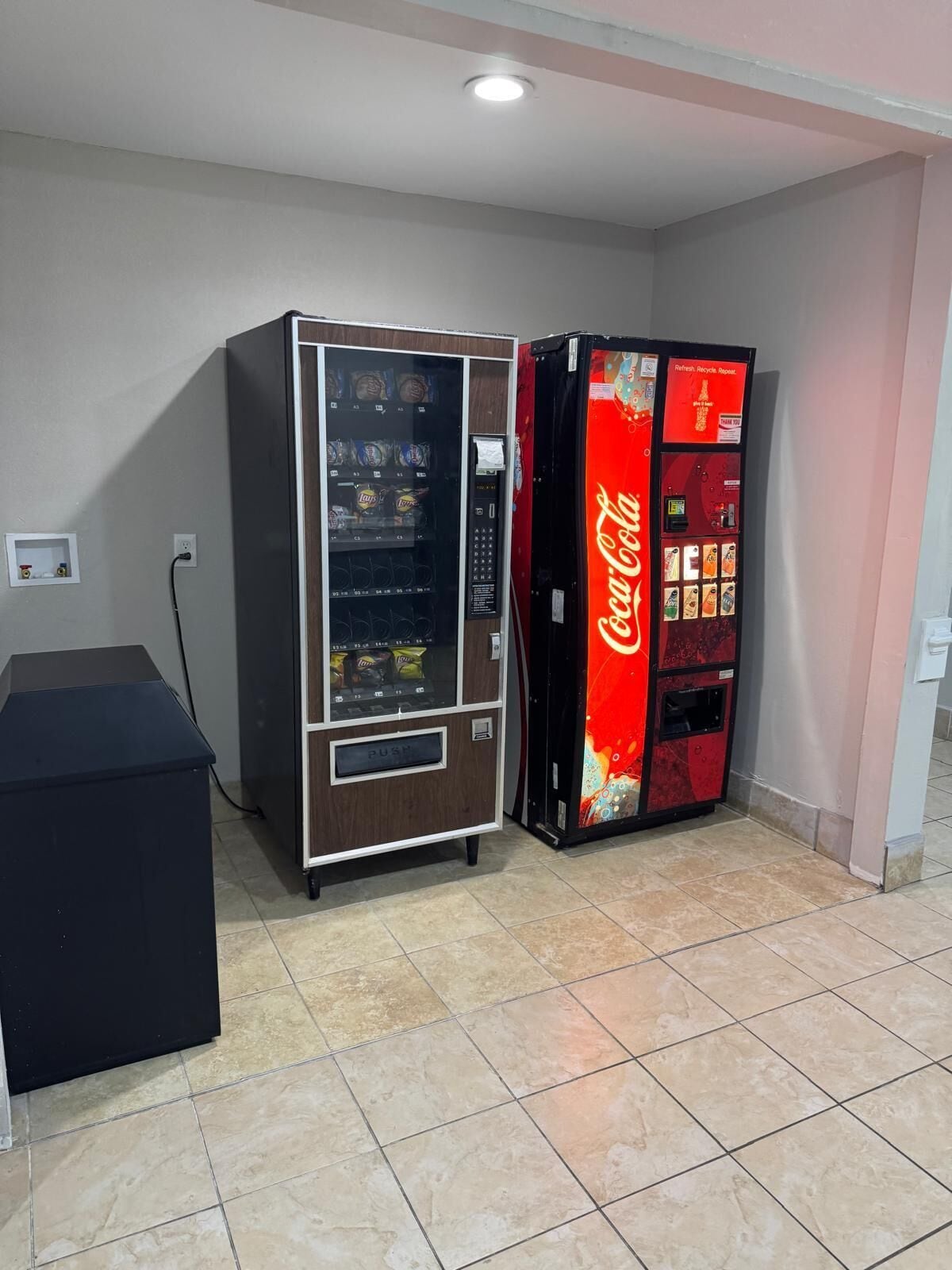 Dues màquines expenedores, una amb la marca Coca-Cola i una altra de color negre, en una sala amb sòl de rajola i sostre blanc.