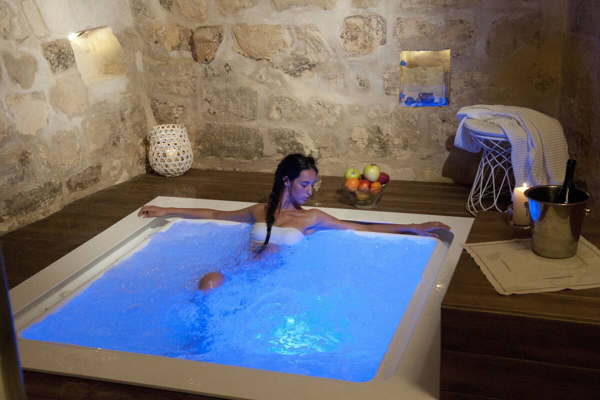 Una persona relajándose en un amplio jacuzzi iluminado, con un fondo de pared de piedra, un jarrón decorativo y un cuenco de frutas sobre una mesa de madera.