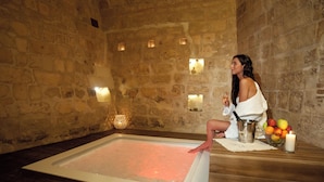A stone-walled room with a large rectangular hot tub, a candle, and a fruit bowl on a wooden table. - MATA SUITE (Matera)