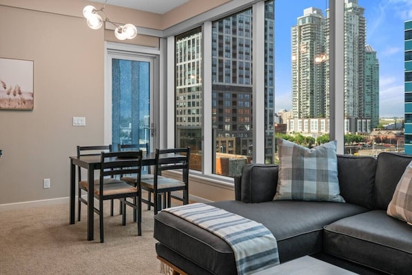 Uma sala de estar moderna com uma mesa de jantar, um sofá com manta e amplas janelas com vista para a cidade.