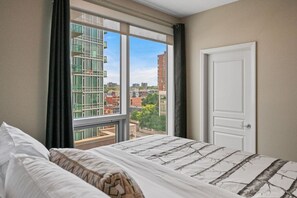 Quarto de hotel com uma janela grande, cama com roupa de cama estampada, uma porta e vista para a paisagem urbana.