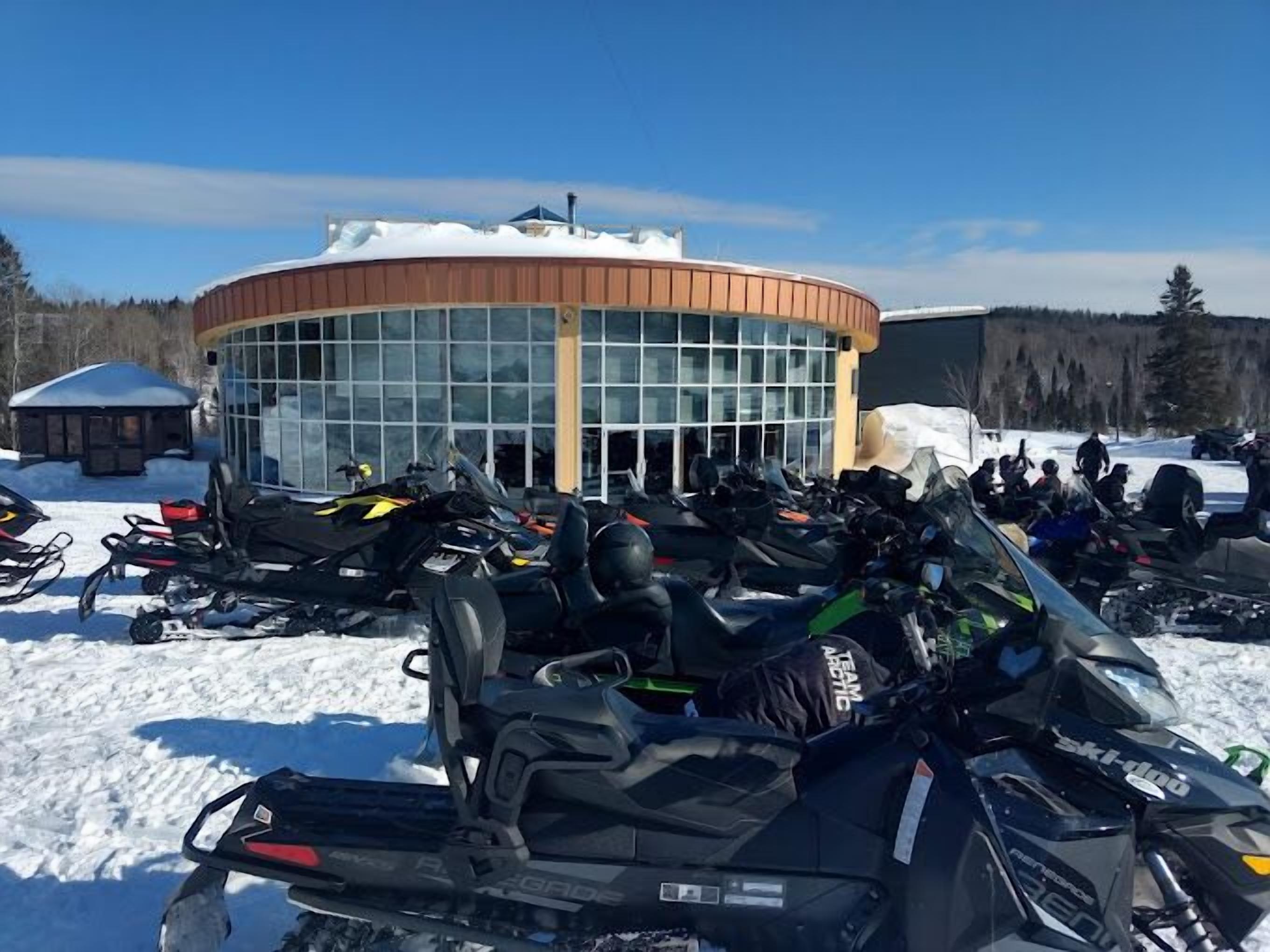 A snow-covered outdoor area with a circular building, possibly a lodge or restaurant, and several snowmobiles parked in front.