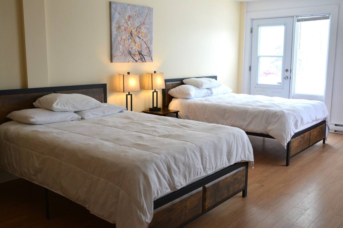 Habitación con dos camas, cada una con ropa blanca y cabeceros de madera. Hay dos mesitas de noche con lámparas, y se observa un cuadro en la pared.