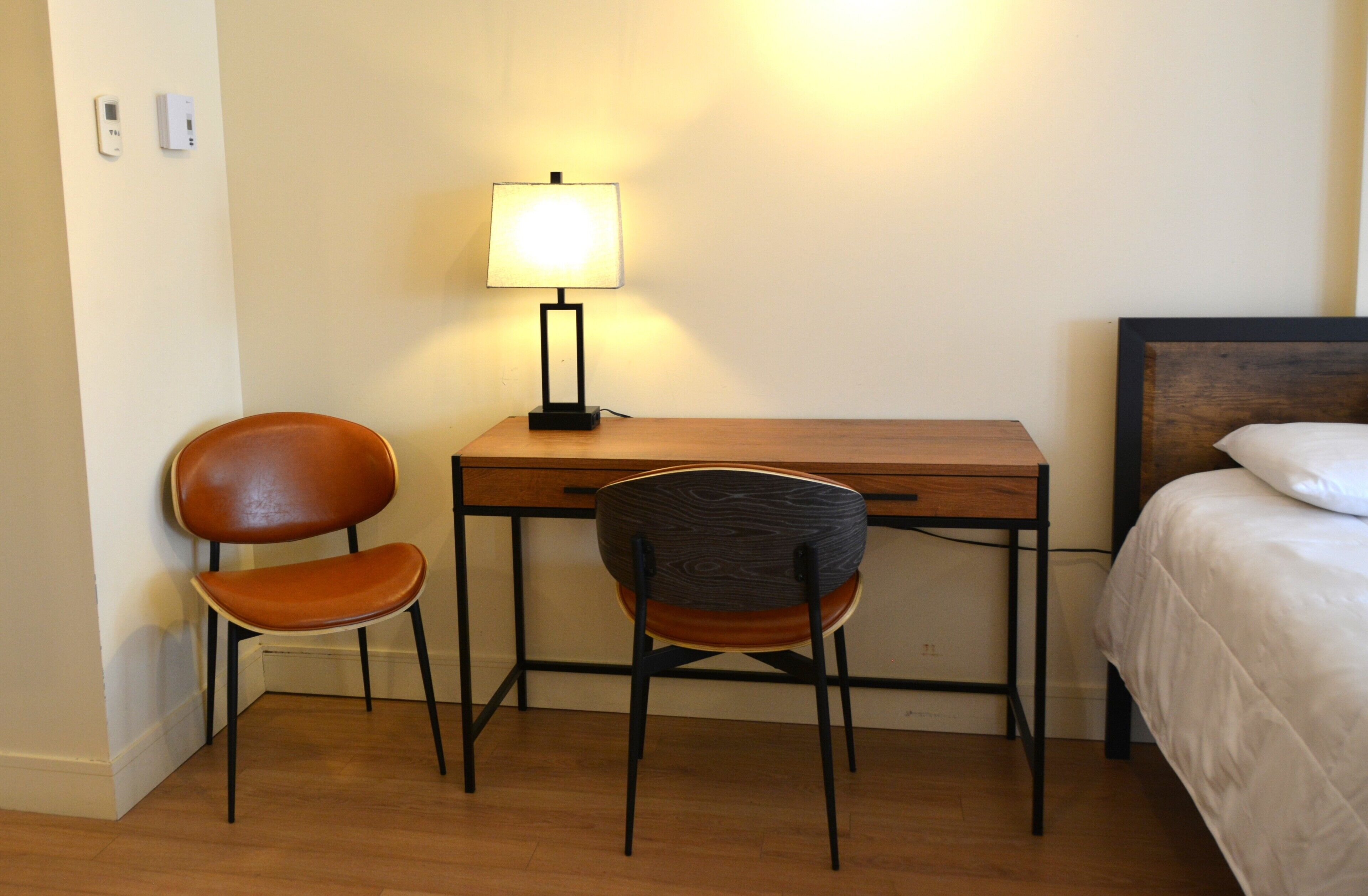 Une petite pièce avec un bureau en bois, deux chaises, une lampe, un lit et un thermostat fixé au mur.