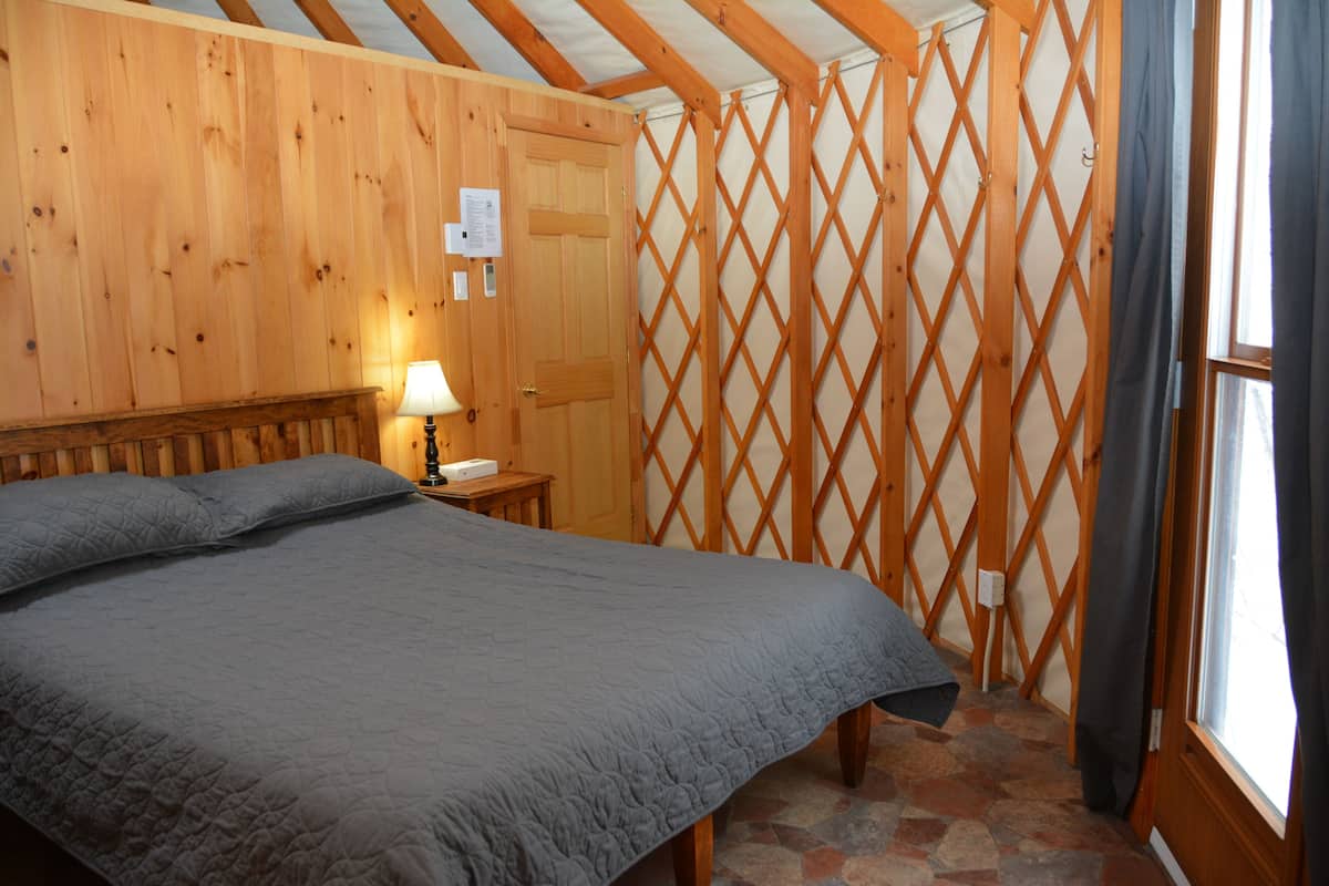 Una habitación con una cama grande, una mesita de noche con lámpara, paredes de madera, un separador de celosías y una ventana con cortinas.