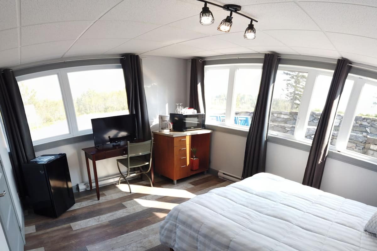 Habitación de hotel con cama, escritorio con computadora, televisor y cómoda. Hay ventanas con cortinas y vistas al exterior.