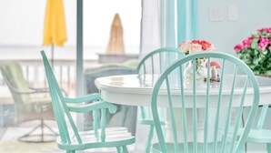 Ein türkisfarbener Holztisch mit Stühlen, eine Vase mit rosa Blumen und Blick auf das Meer durch ein Fenster.