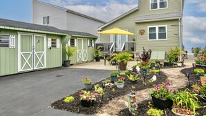 Ein zweistöckiges Gebäude mit grüner Fassade, einer überdachten Terrasse mit gelbem Sonnenschirm und einem Garten mit verschiedenen Topfpflanzen.