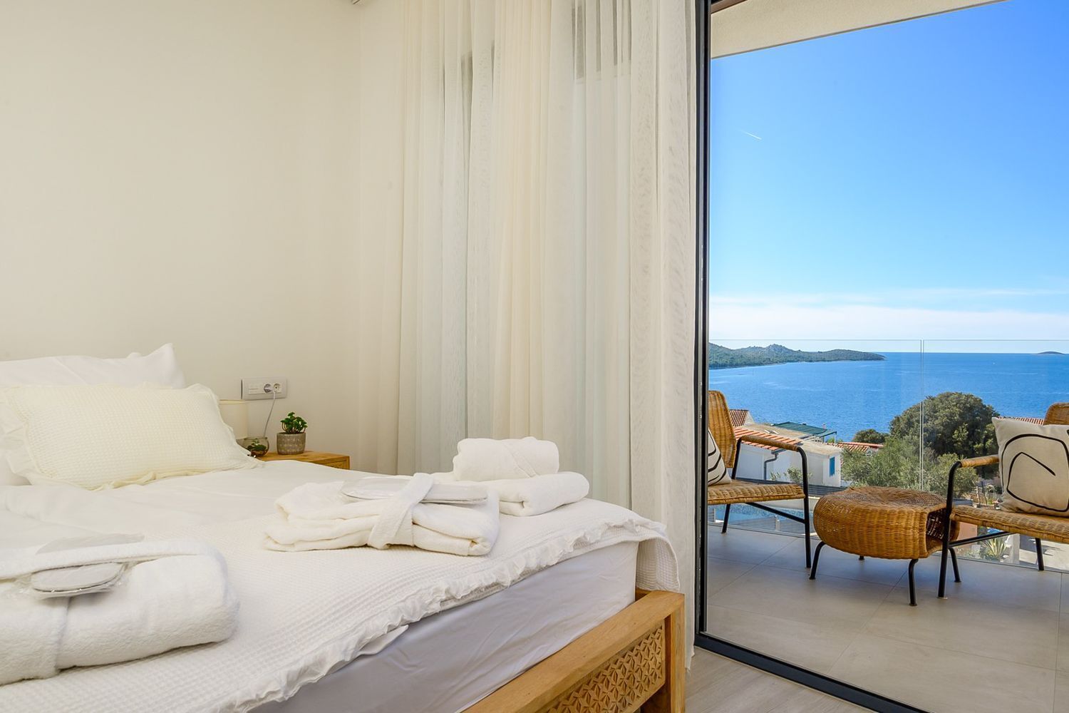 A bedroom with a large bed, white linens, a nightstand with a lamp, and a view of the sea through a glass door.