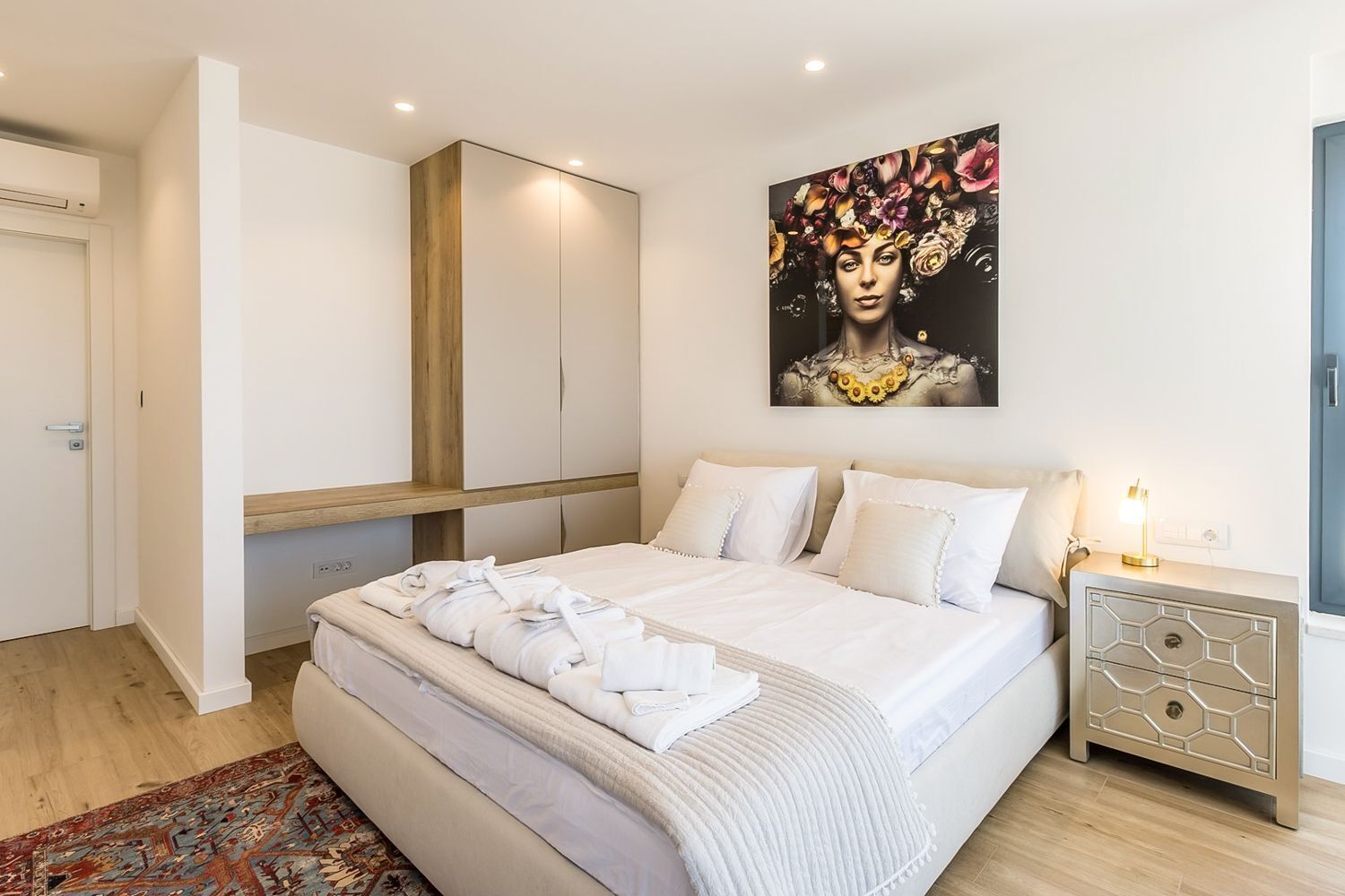 A modern bedroom with a large bed, a nightstand, a wooden wardrobe, and a wall-mounted painting.