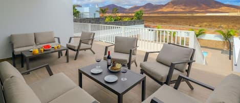 A terrace with outdoor furniture, a table set with plates, glasses, and a bottle, and a view of mountains in the background.