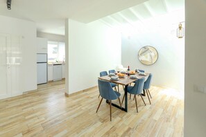 Une cuisine moderne avec des meubles blancs, un coin repas doté d’une table en bois et de chaises bleues, et une grande fenêtre laissant entrer la lumière naturelle.