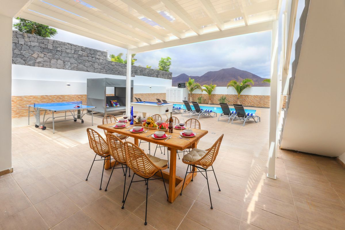 An outdoor patio with a dining table set for four, surrounded by wicker chairs. A pool area with lounge chairs and a barbecue grill is visible in the background.