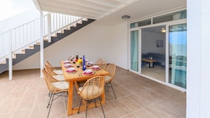 Een eetruimte met een houten tafel gedekt voor vier personen, omringd door rieten stoelen. Een trap is links zichtbaar, en een schuifdeur leidt naar een buitenruimte.