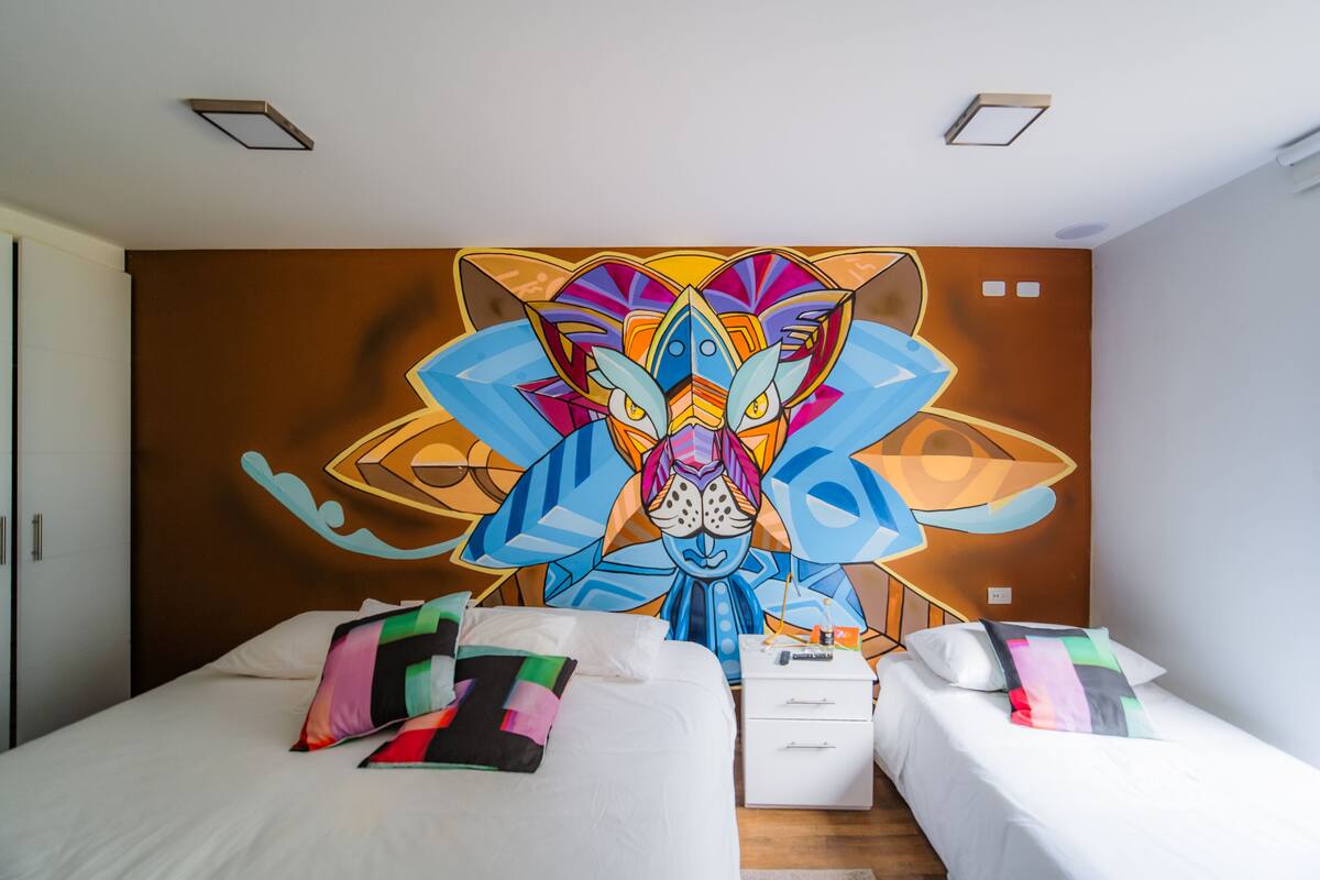 A hotel room with two beds, a colorful mural on the wall, and a small bedside table.