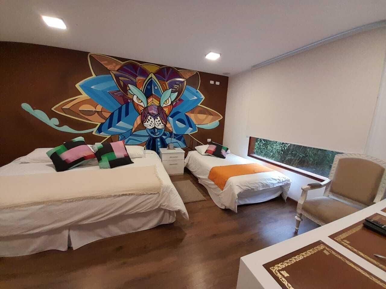 A hotel room with two beds, a large mural on the wall, a window with a view, and a desk with a chair.