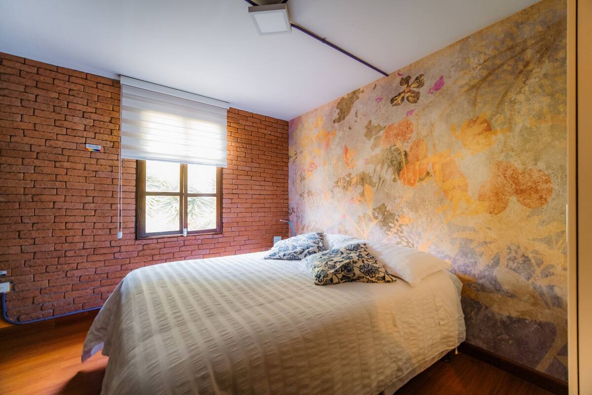 A bedroom with a brick wall, a large window with blinds, a bed with patterned pillows, and a floral wallpaper on the adjacent wall.