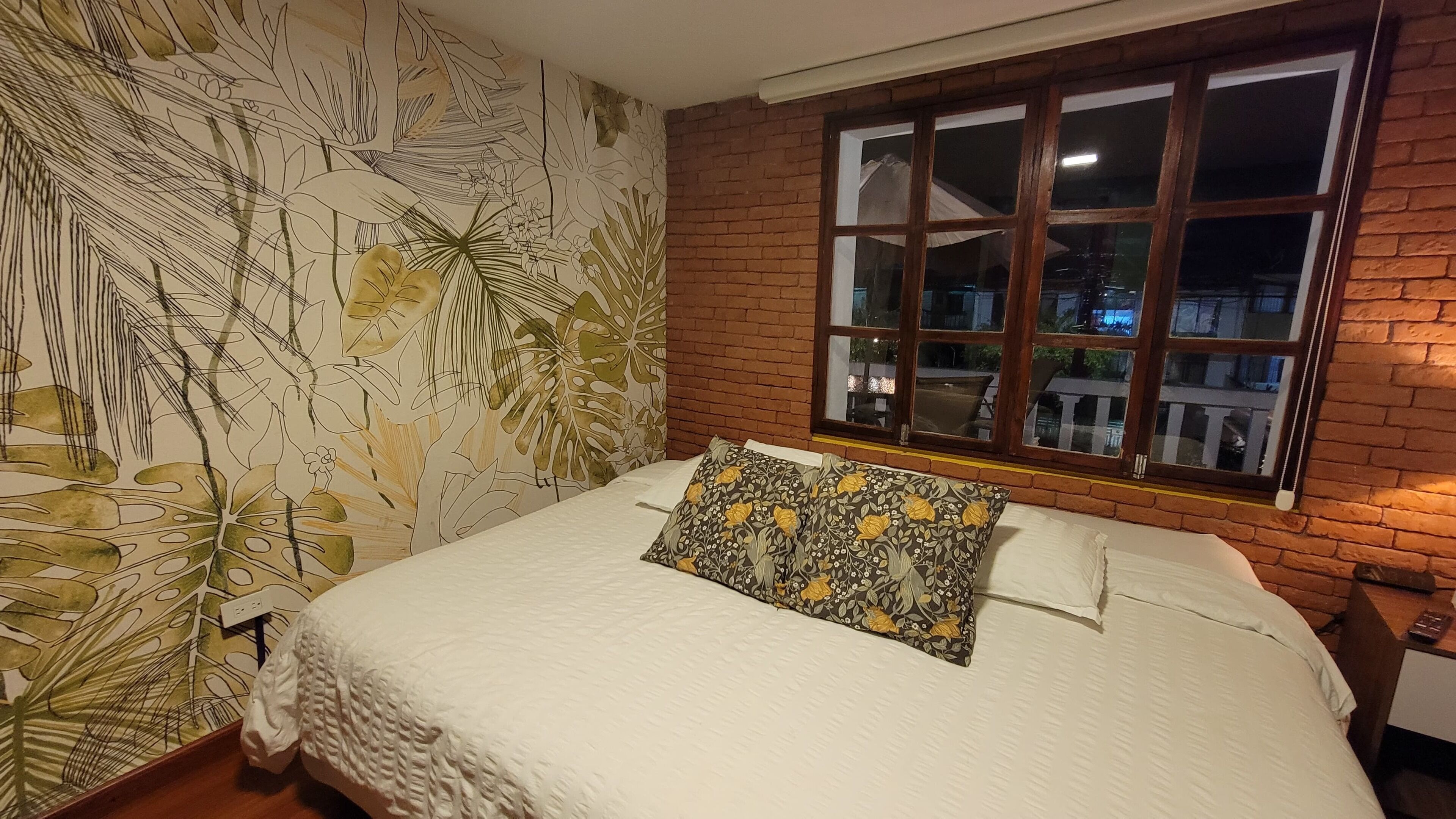 Un dormitorio con pared de ladrillo, una ventana grande con múltiples divisiones y una cama con cubrecama blanco y almohada estampada.