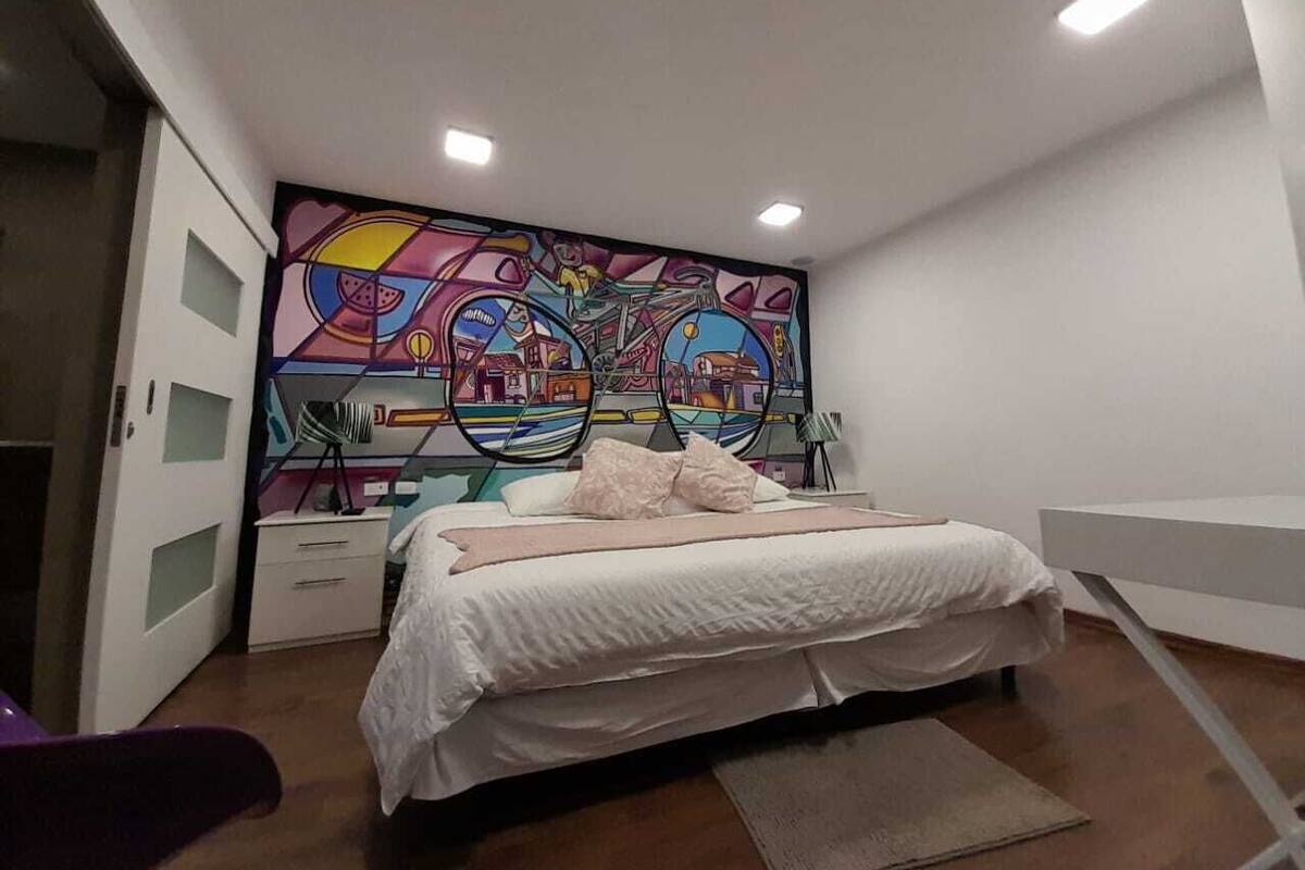 A bedroom with a bed, a nightstand, a dresser, and a colorful abstract mural on the wall.