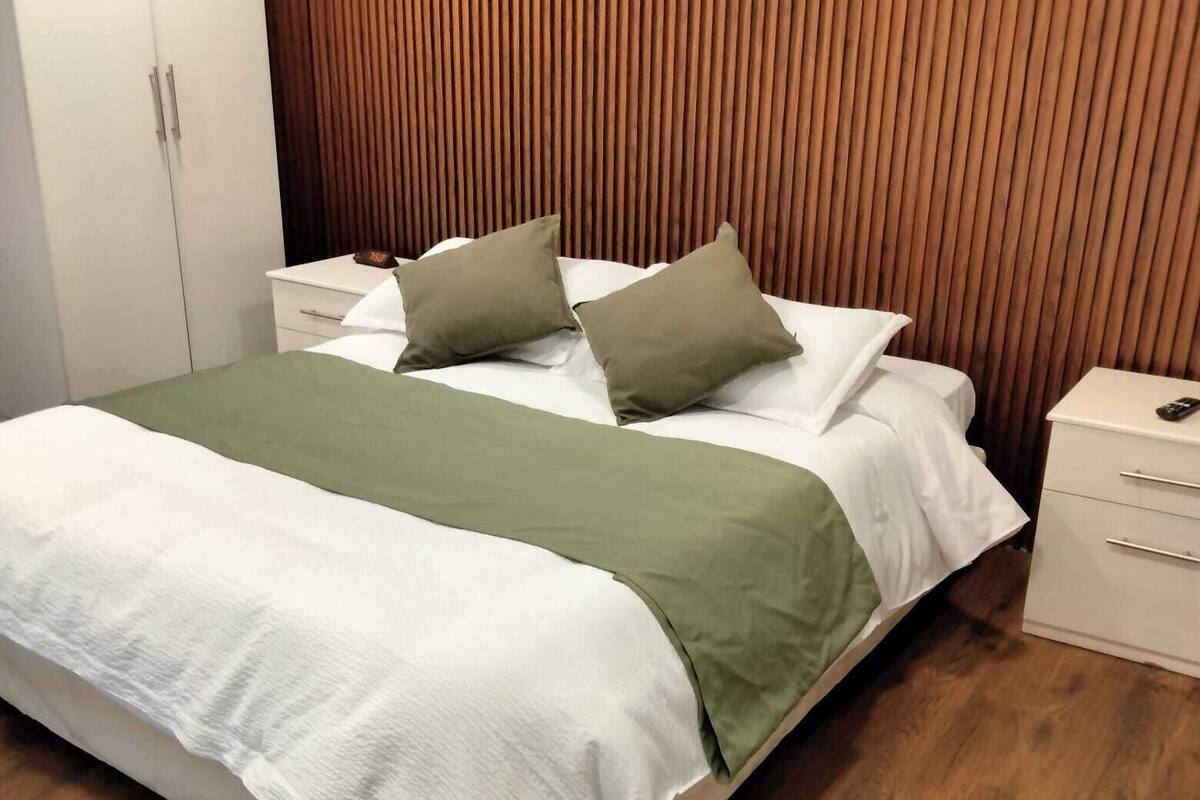A neatly made bed with a green throw blanket and two pillows, a wooden headboard, a white nightstand with a remote control, and a wooden floor.