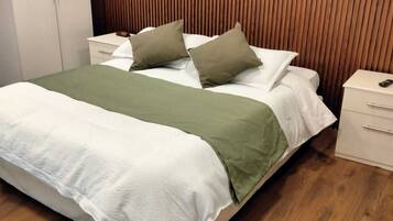 Una cama bien hecha con una manta verde y dos almohadas, un cabecero de madera, una mesita de noche blanca con un control remoto y un suelo de madera.