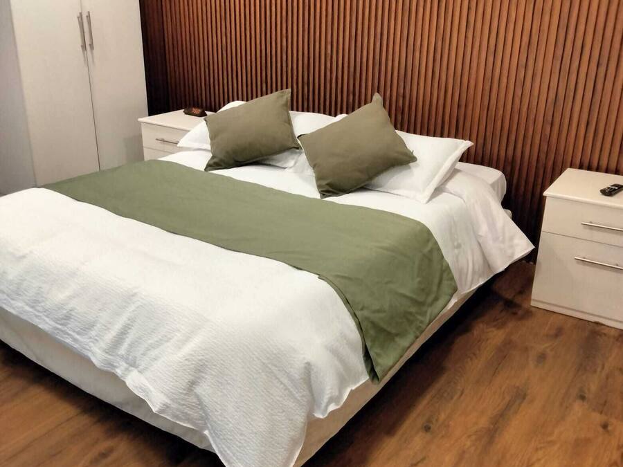 Una cama bien hecha con una manta verde y dos almohadas, un cabecero de madera, una mesita de noche blanca con un control remoto y un suelo de madera.