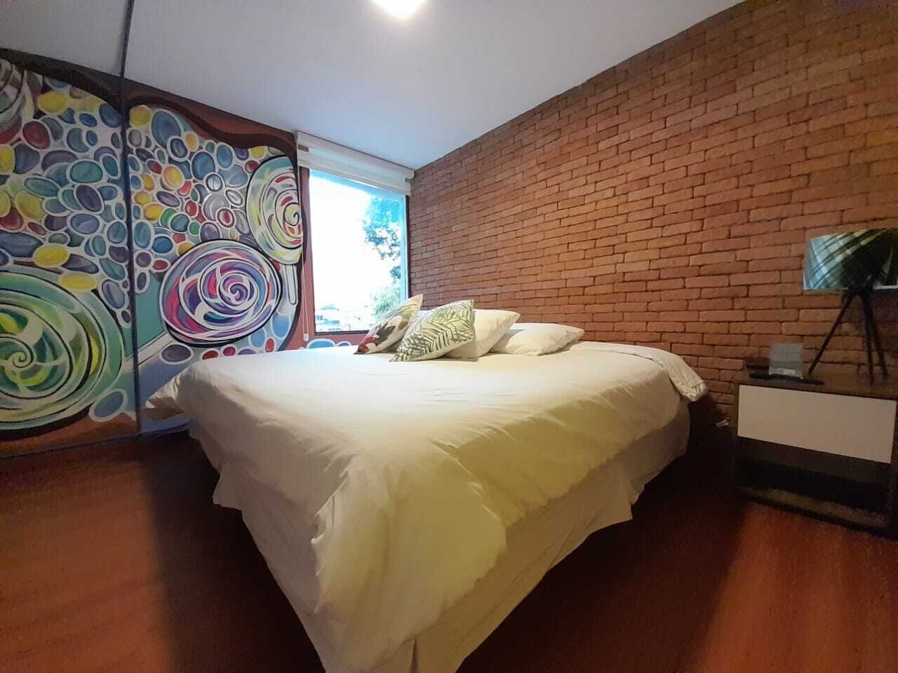 A bedroom with a brick wall, a bed with white bedding, and a colorful abstract painting on the wall.