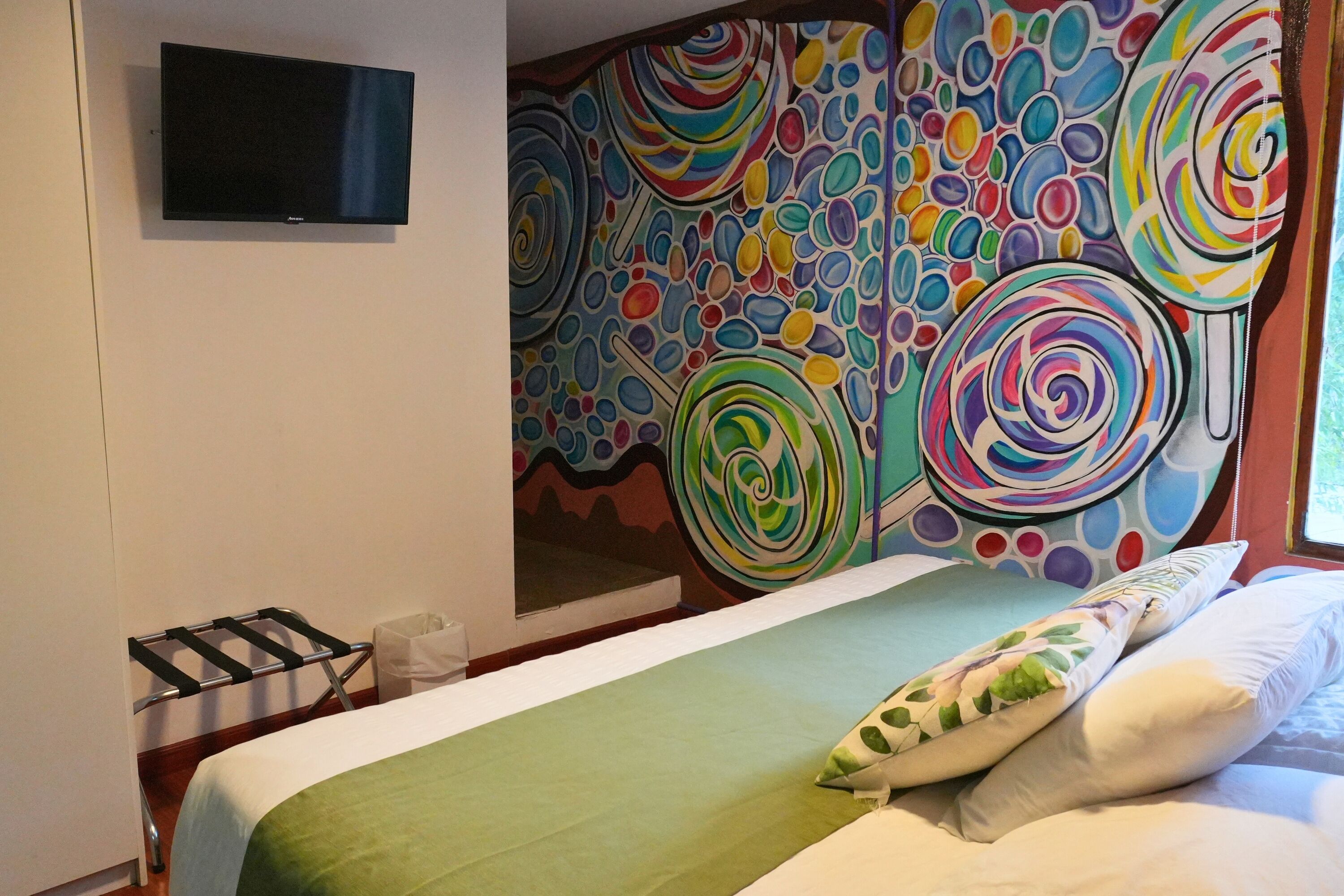 A hotel room with a bed, a TV mounted on the wall, and a colorful abstract mural.