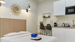 A modern bedroom with a large bed, a wooden headboard, a small kitchenette, and two black chairs. - Charon 4 - Studio Apartment, Sleeps 2 (Paris)