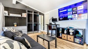 Ein modernes Wohnzimmer mit einem großen Flachbildfernseher, der verschiedene Sender anzeigt, einem dunkelgrauen Sofa mit gemusterten Kissen, einem kleinen schwarzen Tisch und einem Holzboden.