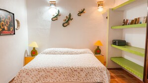 A neatly made bed with a patterned bedspread, two bedside lamps, a wall-mounted shelf with books, and a decorative wall piece. - Standard 1 bedroom suite  #7
 (San Patricio)