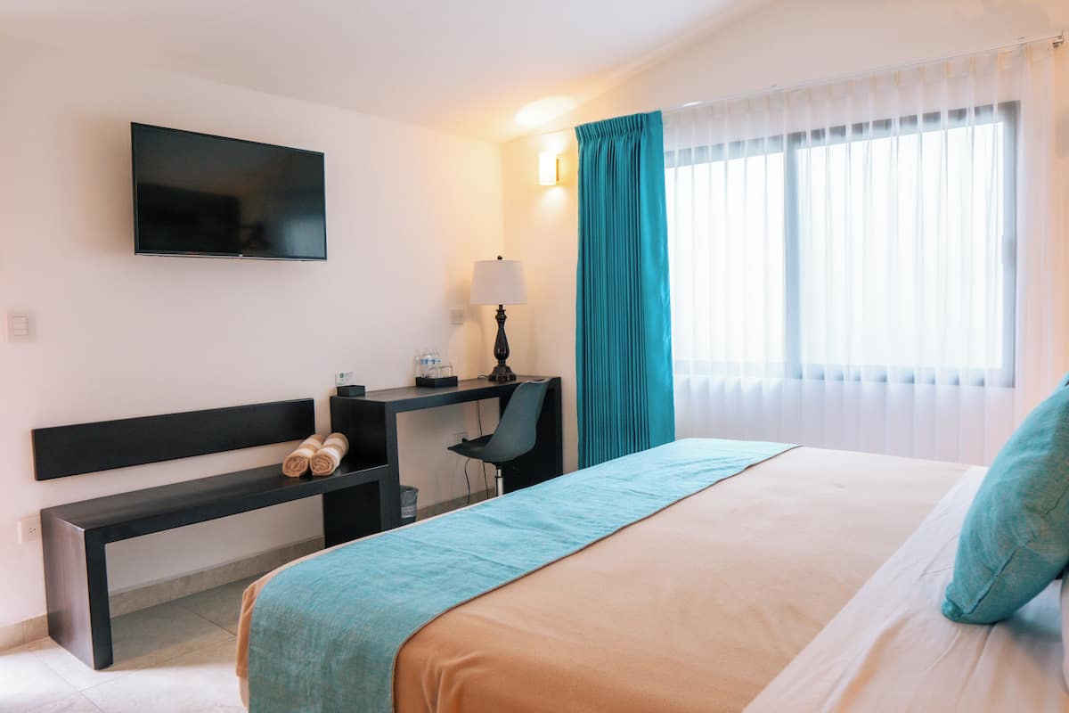 A hotel room with a bed, a desk with a chair, a television mounted on the wall, and a window with curtains.