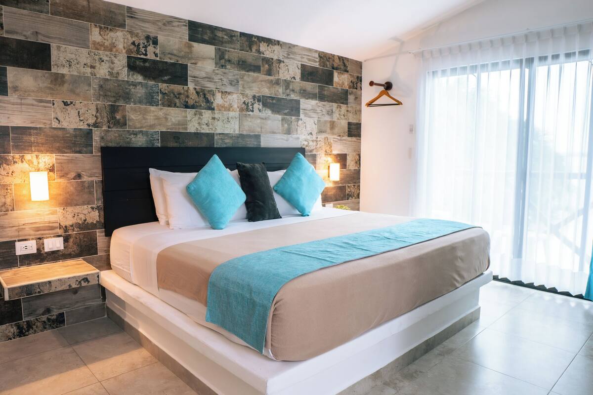 A modern hotel room with a large bed, a wooden accent wall, and a view of the outdoors.
