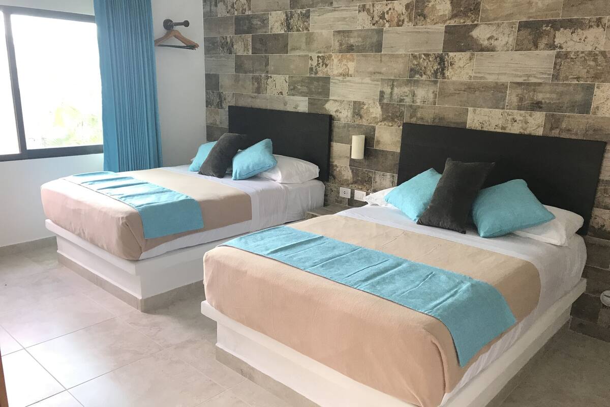 Two beds with beige and blue bedding, a stone-patterned wall, and a window with blue curtains.