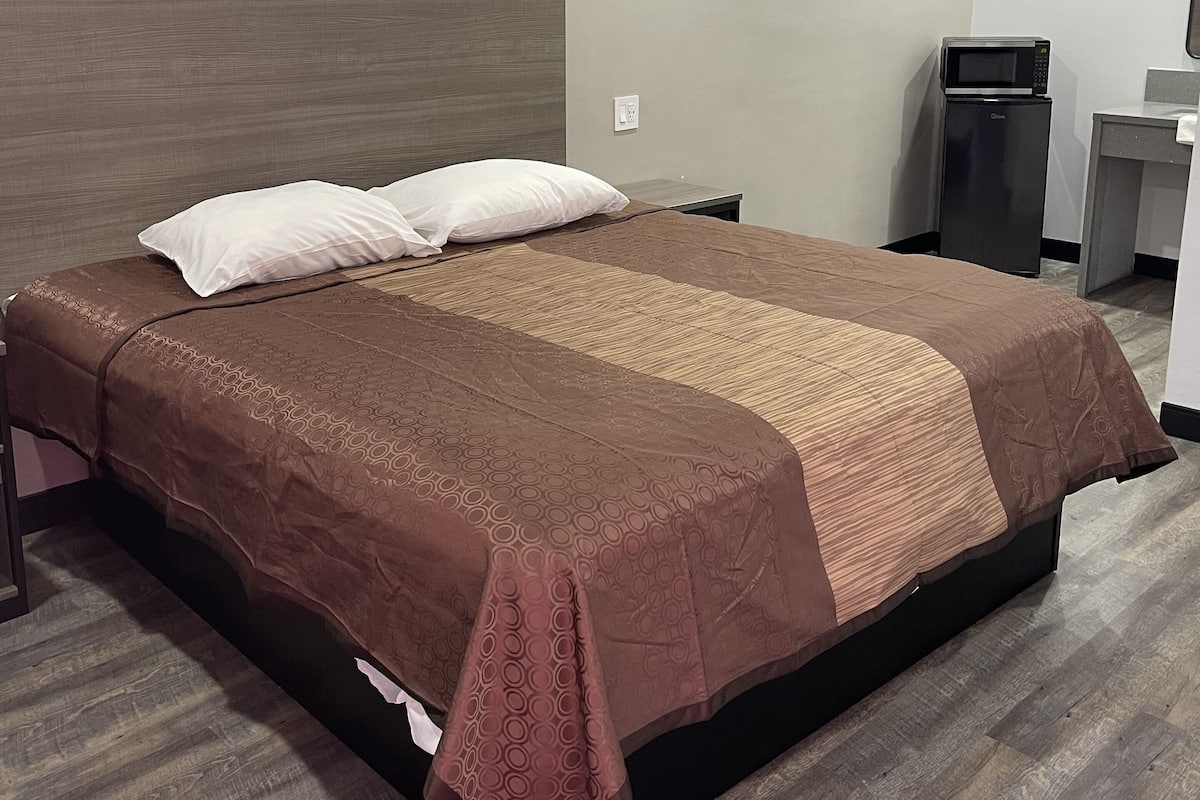 A hotel room with a large bed, a bedside table, a microwave, and a small desk with a chair.