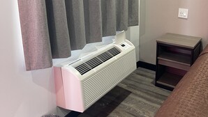 A hotel room with a built-in air conditioning unit, grey curtains, a small bedside table, and a patterned bedspread. - Welcome Inn & Suites (Anaheim)