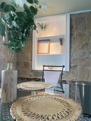 A room with a stone wall, a white shelf with a framed picture, a chair, a table with a woven placemat, and a large plant. - The Whiskey House 12 min from Round Top, steps away from Kooper Family Whiskey (Ledbetter)