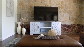 Ein Wohnzimmer mit Steinwand, einem Flachbildfernseher auf einem weißen Schrank, einem braunen Ledersitzhocker und Dekorationselementen.