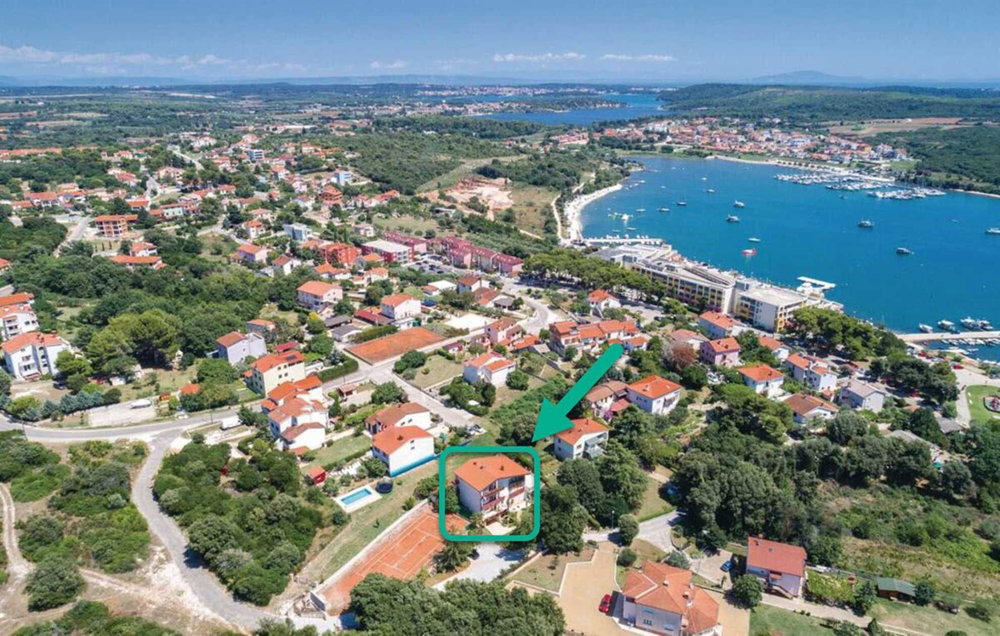 An aerial view of a coastal town with a marina, residential buildings, and a prominent hotel with a red roof.